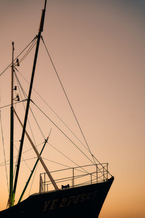 A fishing boat is silhouetted beneath tall masts at sunset, with warm colors filling the sky in a...