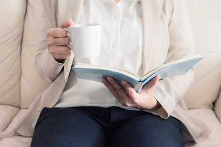 woman is sitting on a sofa and reading a book while holding a coffee cup. retro style photoの写真素材