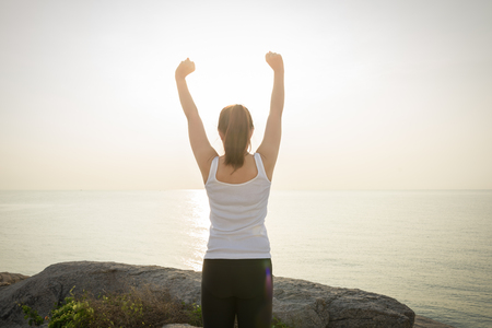 Happy successful sportswoman raising arms to the sky on golden back lighting sunrise summer. Fitness athlete with arms up celebrating goals after sport exercising and working out outdoors background is seaの写真素材