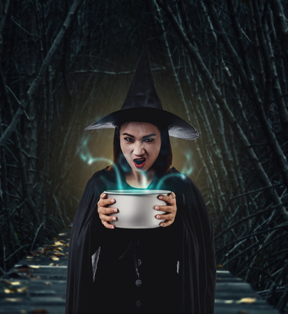 Portrait of woman in black Scary witch halloween costume holding spooky witch's cauldron smoke coming out with moonlight in a dark forestの写真素材