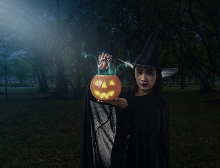 Portrait of woman in black Scary witch halloween costume Holding halloween pumpkin with moonlight in a dark forestの写真素材