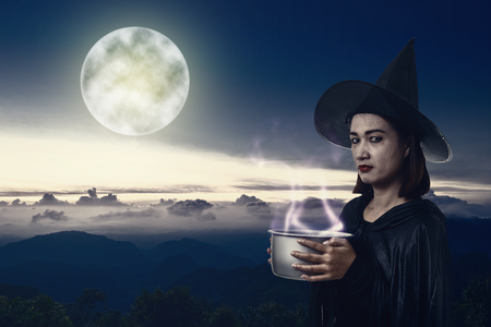 Portrait of woman in black Scary witch halloween costume holding spooky witch's cauldron smoke coming out with moonlight. landscape mountain and sea of mist is backgroundの写真素材