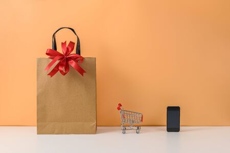 Paper shopping bags and shopping cart or trolley with Red bow and smartphone on white table and pastel orange background. with copy space for your message. The concept of selling or shopping onlineの写真素材