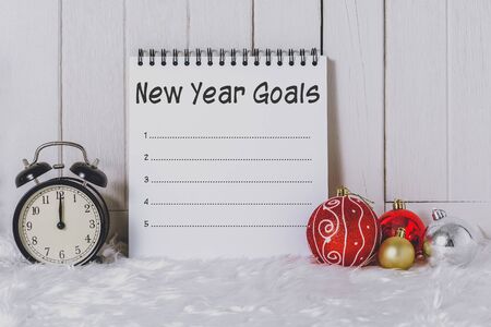Alarm clock with Christmas ornaments and New Year's Goals List written on Notebook with white fur and white wooden backgroundの写真素材