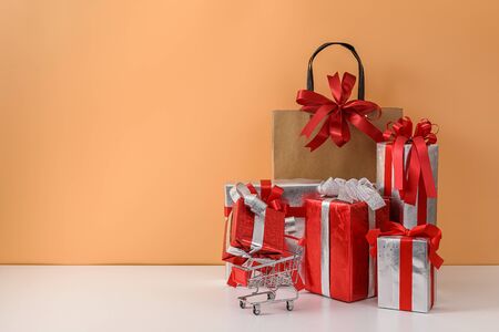 Paper shopping bags and shopping cart, trolley. many Gift Box with Red bow on white table and pastel orange background. with copy space for your message. Shopping New Year's gift, Christmas Conceptの写真素材