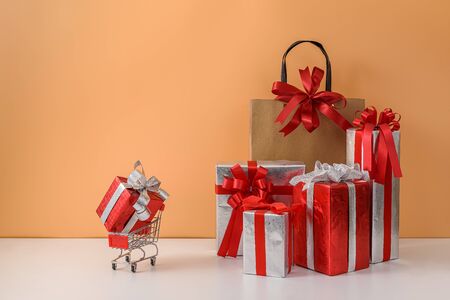 Paper shopping bags and shopping cart, trolley. many Gift Box with Red bow on white table and pastel orange background. with copy space for your message. Shopping New Year's gift, Christmas Conceptの写真素材
