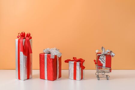 Many Gift Box with Red bow on shopping cart, trolley on white table and pastel orange background. with copy space for your message. The concept of selling shopping online, New Year's gift or Christmasの写真素材
