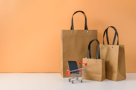 Paper shopping bags and shopping cart or trolley with smartphone on white table and pastel orange background. with copy space for your message and logo. The concept of selling or shopping onlineの写真素材