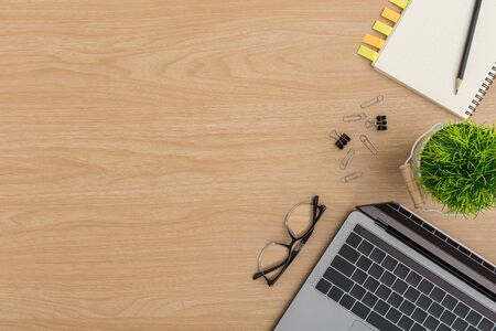 Top view Wood office desk table. Flat lay Workspace with eye glasses, tree pot, laptop, paper clip, clipboard, notebook, pencil, sticky note office supplies on wooden backgroundの写真素材
