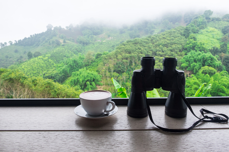 binoculars and coffee cup on wooden with forest on the hill blurred background.の写真素材