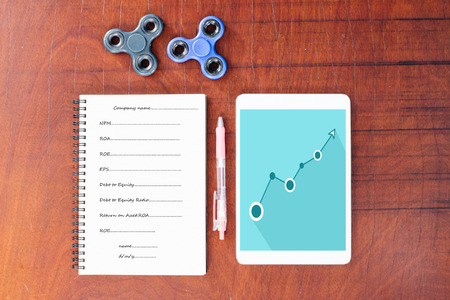 Blue graph on tablet, Pen, Black and blue hand spinner at the top notebook on wood table background, The notebook had vocabulary about business.の写真素材