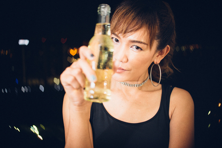 Happy asian woman in black dress looking you while holding beer bottle, background is pub have bokehの写真素材