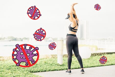 Asian woman doing stretching Exercise and Social distancing protect Covid-19 and other Contagious disease at outdoors along city sidewalk and listening to music with earphones  in summer warm lightの写真素材
