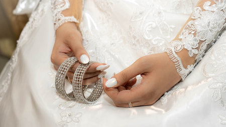 Detail of a bride's hands holding her bangles with bridal white dress and white nails and an...
