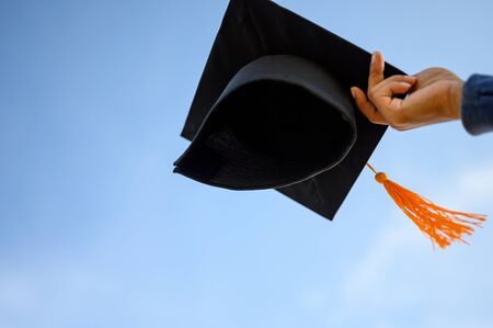 Graduates hold a black hat with a yellow tassel attached to the sky.の写真素材