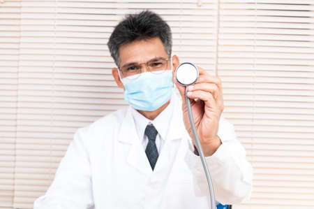 A male doctor, wearing a mask and white adhesive clothes, sits at his desk.の写真素材