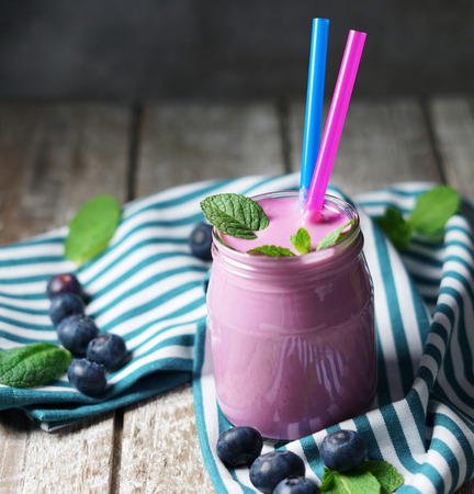 Blueberry fresh smoothie milkshake in a glass with two drinking straw and mint leaves on black rustic background and a wooden table.の写真素材