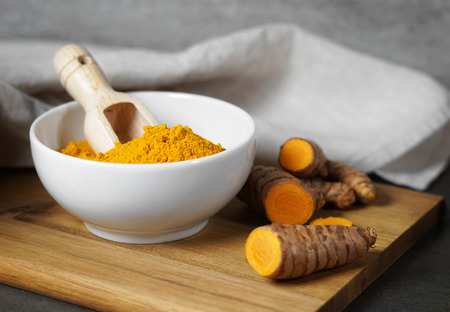 Turmeric powder and sliced turmeric roots healthy spice Asian food closeup of a white bowl with a wooden scoop with copy space.の写真素材