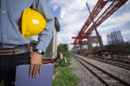 Engineer holding yellow helmet for workers security on background of construction cranes.