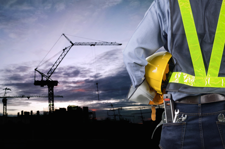 Asian engineer holding yellow helmet for workers security on background of new highrise apartment buildings and construction cranes on background of evening sunset Silhouette Crane lifts load.の写真素材