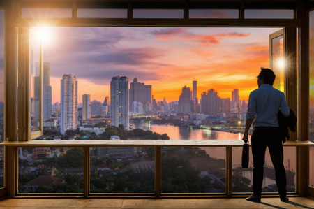 Concept vision, Young businessman wearing comfortable casual suit jacket standing holding a work bag in Working room near the glass window and looking through the modern buildings in the skyline.の写真素材
