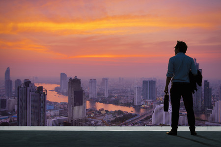 Concept vision, Young businessman wearing comfortable casual suit jacket standing holding a work bag on top of building and looking through the modern buildings in the skyline.の写真素材