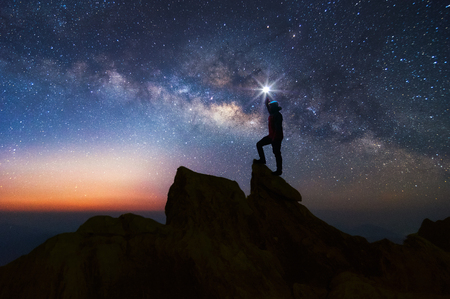 Silhouette of climber or backpacker. In his hand, he held the light up high above his head. Standing on rocky mountain peak and Milky Way Galaxy, success, winner, leader concept. High iso with Noise.の写真素材