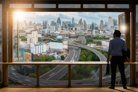 Concept vision, Young businessman wearing comfortable casual suit jacket standing holding a work bag in Working room near the glass window and looking through the modern buildings in the skyline.の写真素材