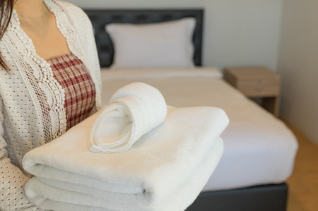 Young hotel maid holds a towel, sheets and soft towels in the clean room. The hotel has a clean white blanket on the bed with a pillow in the morning bedroom.の写真素材