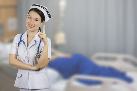 Asian beautiful young nurse in white uniform with background blur patient on bed in room hospital.の写真素材