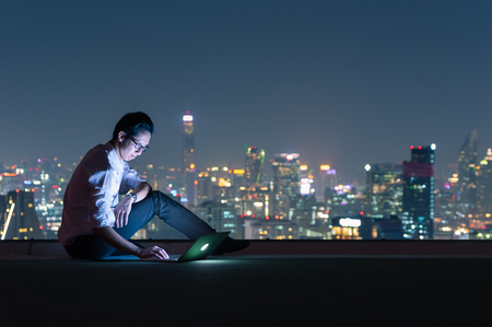 Asian businessman sitting at the top of the building. Use the laptop computer with the scenery of the night view of the city, Business success and technology concept.の写真素材