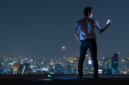 Asian businessman stand at the top of the building. Use the smartphone and laptop computer with the scenery of the night view of the city, Business success and technology concept.の写真素材