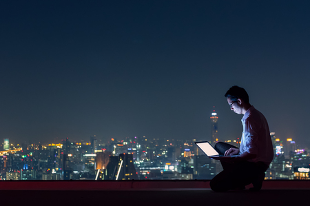 Asian businessman sitting at the top of the building. Use the laptop computer with the scenery of the night view of the city, Business success and technology concept.の写真素材