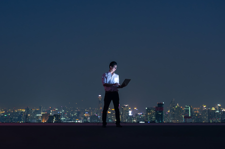 Asian businessmen stand at the top of the building. Use the laptop, computer with the scenery of the night view of the city, Business success and technology concept.の写真素材