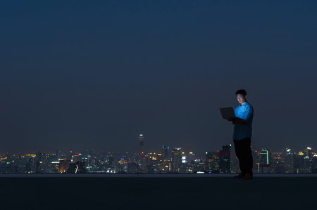 Asian businessmen stand at the top of the building. Use the laptop, computer with the scenery of the night view of the city, Business success and technology concept.の写真素材