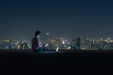 Asian businessman sitting at the top of the building. Use the smartphone and laptop computer with the scenery of the night view of the city, Business success and technology concept.の写真素材