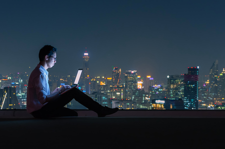 Asian businessman sitting at the top of the building. Use the laptop computer with the scenery of the night view of the city, Business success and technology concept.の写真素材