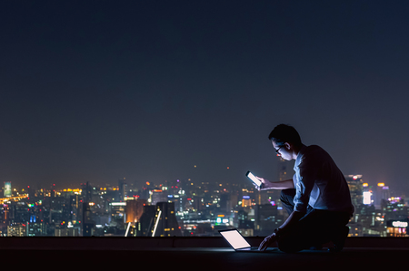 Asian businessman sitting at the top of the building. Use the smartphone and laptop computer with the scenery of the night view of the city, Business success and technology concept.の写真素材