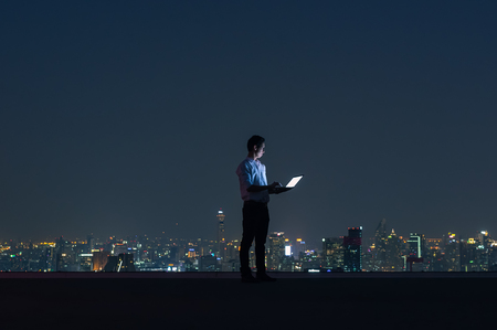 Asian businessmen stand at the top of the building. Use the laptop, computer with the scenery of the night view of the city, Business success and technology concept.の写真素材