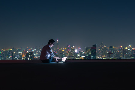 Asian businessman sitting at the top of the building. Use the laptop computer with the scenery of the night view of the city, Business success and technology concept.の写真素材
