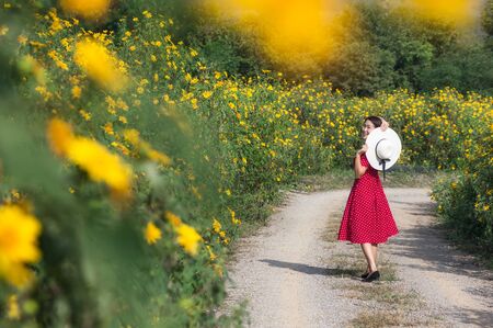 Beautiful woman in red dress and white hat in nature, in rural road, standing back, spring, on Tung Bua Tong Mexican sunflower field in Mae Moh Coal Mine, Lampang Province, Thailand.の写真素材