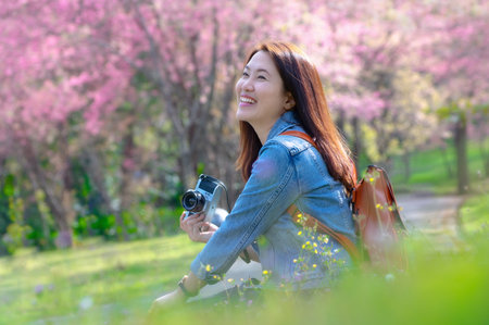 Beautiful young Asian backpacker using vintage camera and laughing and smiling happily in the midst of spring cherry blossoms bloom Travel lifestyle, world explorer, or summer Asia travel idea.の写真素材