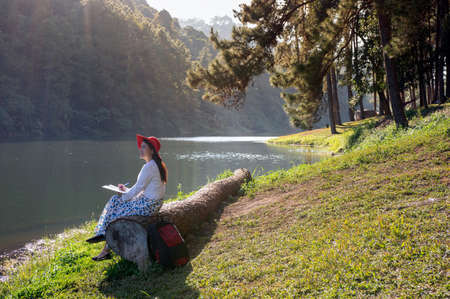 Beautiful asian woman smiling Sit on a large log and write a diary in a lush garden by a shady lake. Pang Oung, Mae Hong Son Province, Thailand.の写真素材