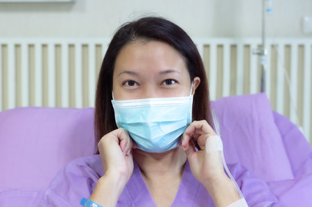 Young Asian female patient is smiling and showing medical mask. Patient feels happy and comfortable with treatment and therapy on hospital bed in hospital room. Medical healthcare concept.の写真素材