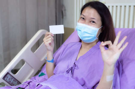 Young Asian female patient is smiling and showing blank credit card, Patient feels happy and comfortable with treatment and therapy on hospital bed in hospital room. Healthcare and Insurance concept.の写真素材