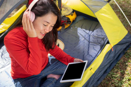 A beautiful woman in a red dress Enjoy listening to music from headphones and use tablet blank. She sat in a yellow tent. Admiring the beautiful nature backpacking camping In the middle of a beautiful pine forest beside Lake, Pang Oung ,Mae Hong Son, Thailandの写真素材