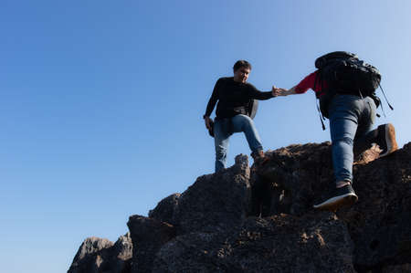 Young man and women backpacker are climbing a mountain, A man stretching his hand to pull a woman up to succeed in their climb, Couple are good teamwork. They both hiked on a clear day.の写真素材