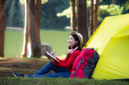 A young girl sits next to a camping tent and a red backpack, sitting on a travel journal and admiring nature in the middle of a pine forest beside the lake, Pang Oung, Mae Hong Son, Thailand.の写真素材