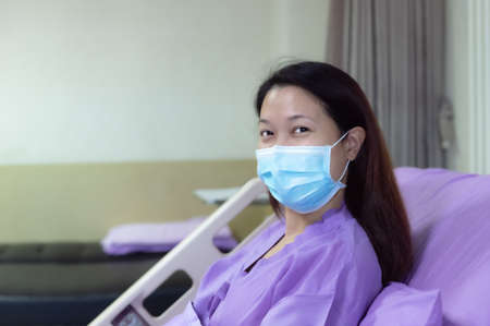 Young Asian female patient is smiling and showing medical mask. Patient feels happy and comfortable with treatment and therapy on hospital bed in hospital room. Medical healthcare concept.の写真素材