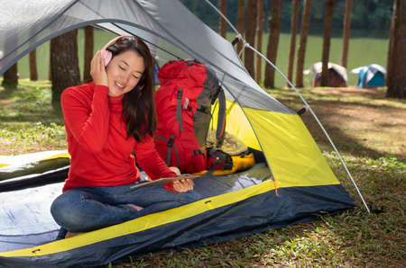 A woman Enjoy listening to music from headphones with play tablet. She sat in a yellow tent. She backpacking camping In the middle of a pine forest beside Lake. Pang Oung, Mae Hong Son, Thailandの写真素材